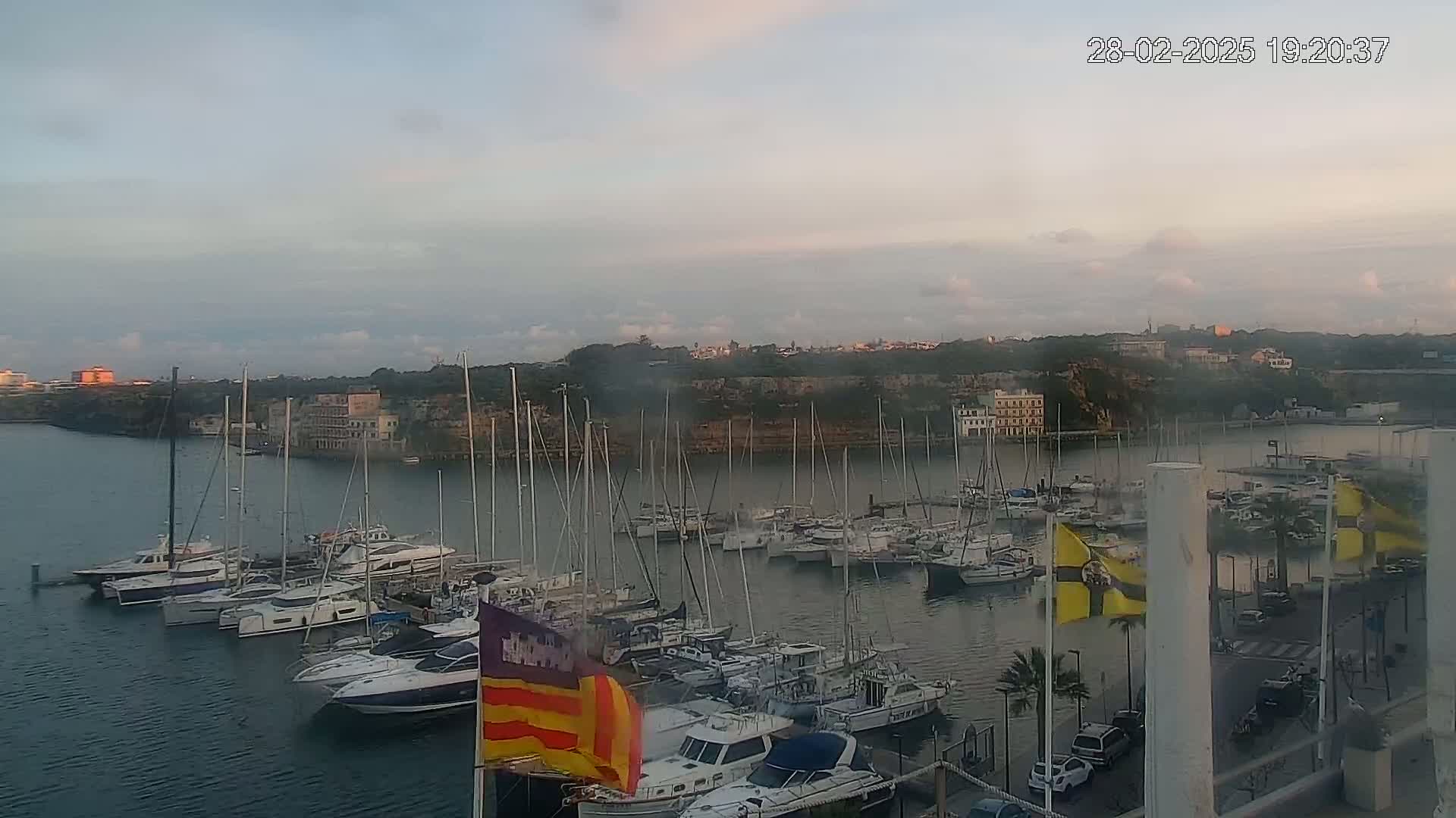 Many sailboats and motorboats are docked in a calm harbor at dusk under a mostly cloudy sky.