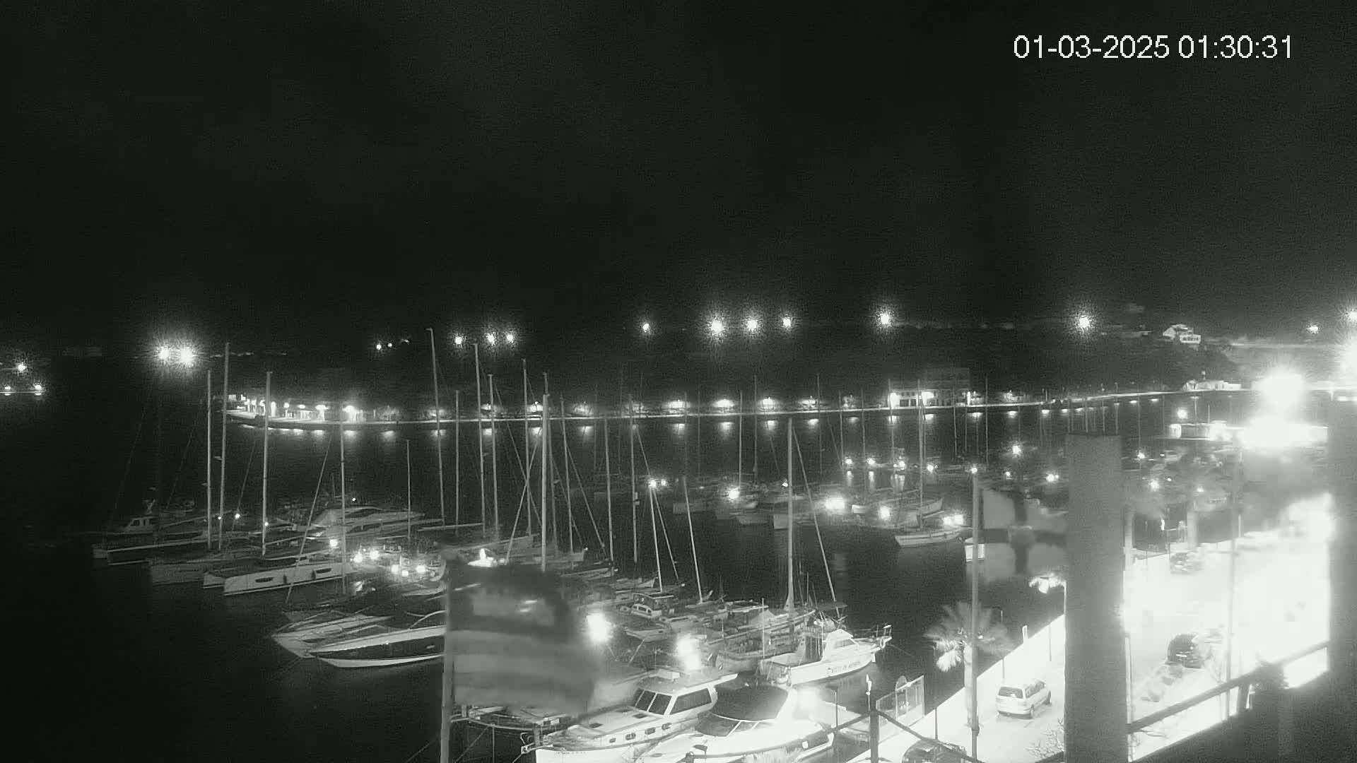 A nighttime, black and white view of a marina filled with docked sailboats and motorboats under a clear sky.