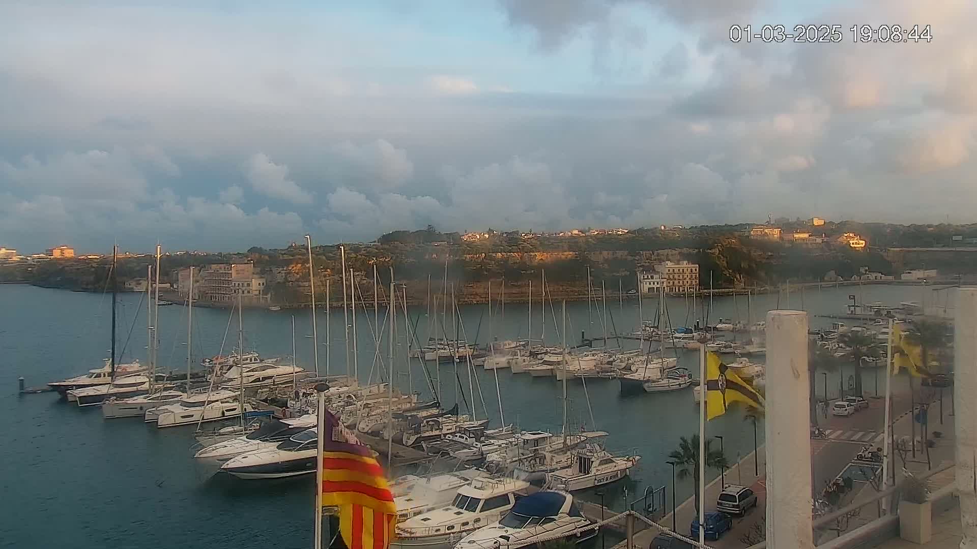 A marina filled with numerous sailboats and motorboats is situated in a calm bay under a partly cloudy sky at sunset.