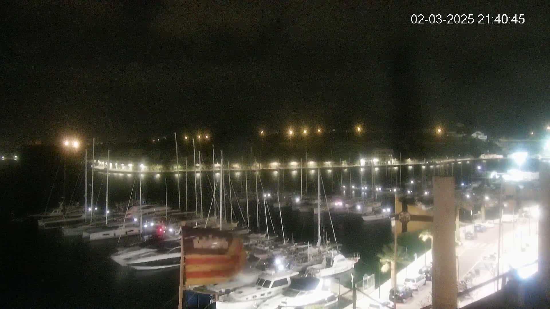 A nighttime view of a marina filled with numerous boats and yachts, illuminated by lights along a waterfront walkway under a clear, dark sky.