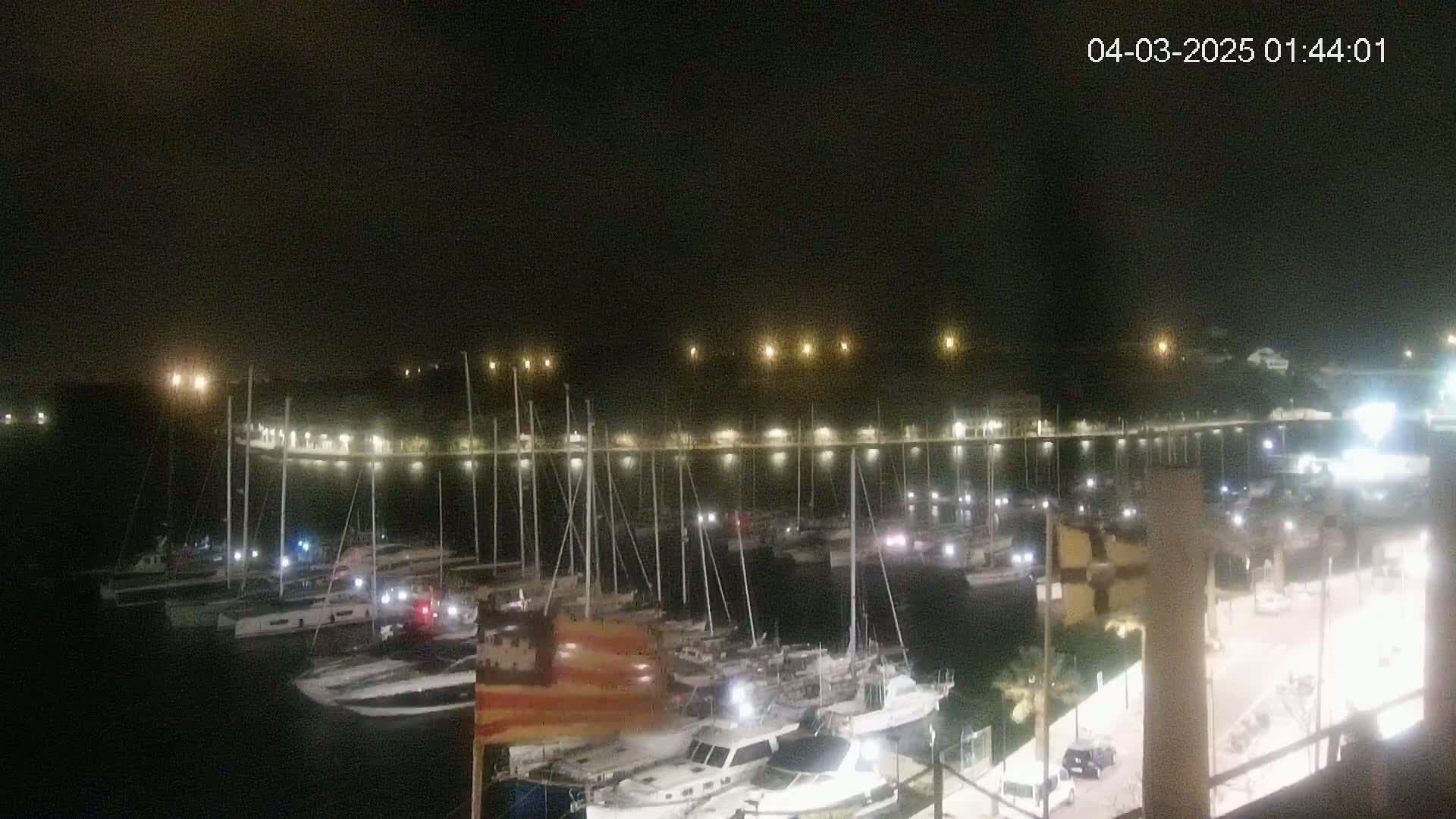 A nighttime view of a marina filled with numerous boats and yachts, illuminated by lights along a waterfront.