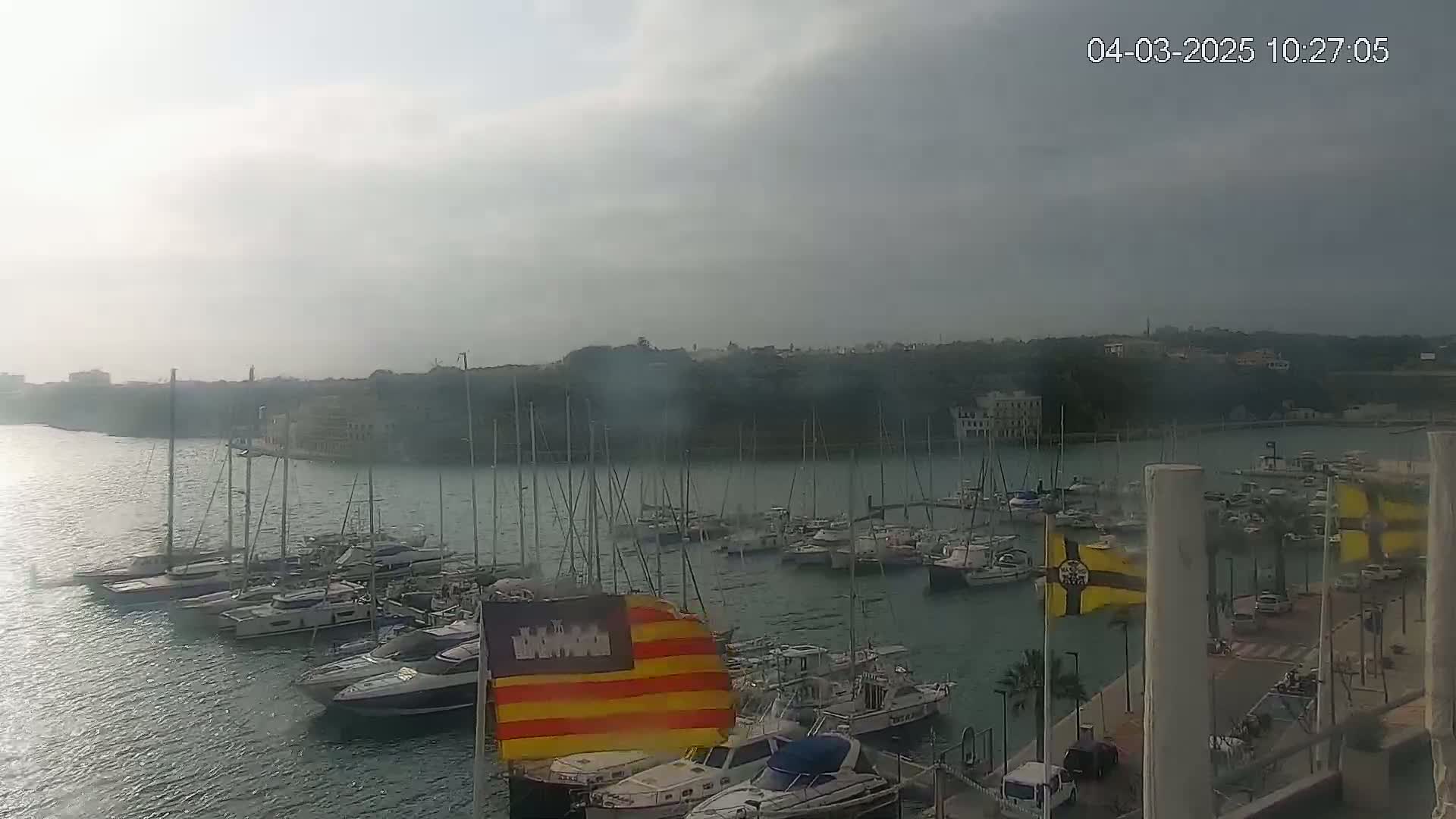 A marina filled with numerous sailboats and motorboats is visible under an overcast sky.