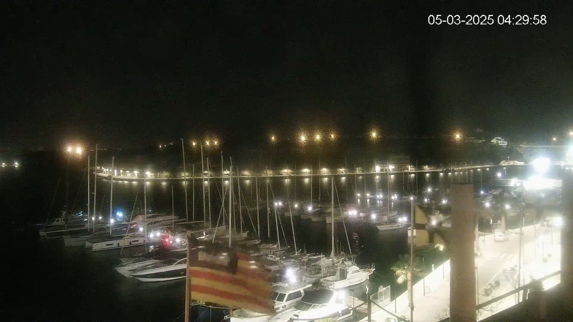A nighttime view of a marina filled with numerous boats and yachts illuminated by lights along the docks and shoreline under a clear, dark sky.