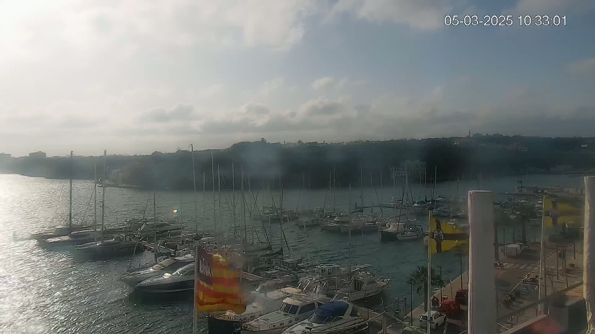 A partly cloudy day reveals a marina filled with numerous sailboats and motorboats docked along a shoreline, with a hilly landscape in the background.