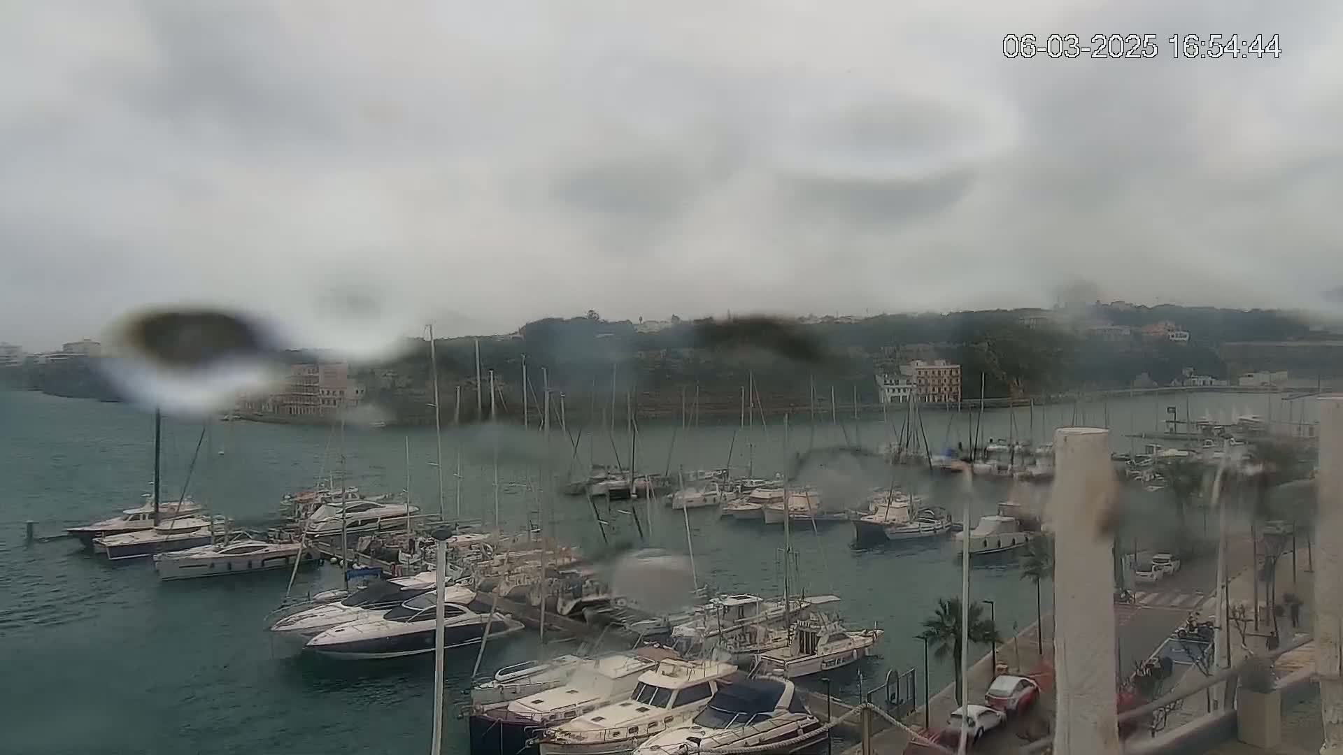 A rain-streaked view of a marina filled with numerous boats and yachts, situated in front of a hillside town under a cloudy sky.