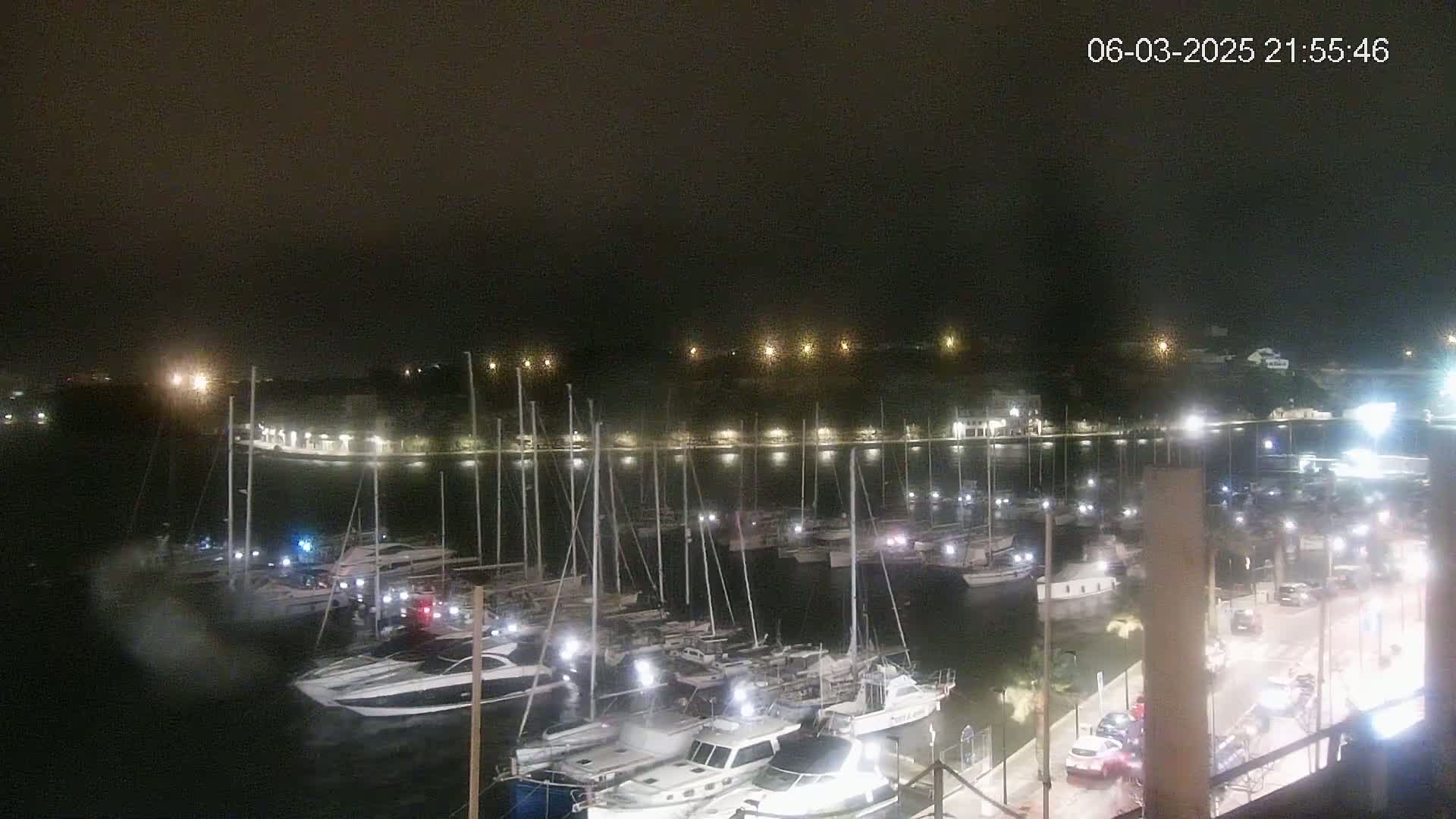 A nighttime view of a marina filled with boats, docked along a waterfront illuminated by lights, under a dark sky.