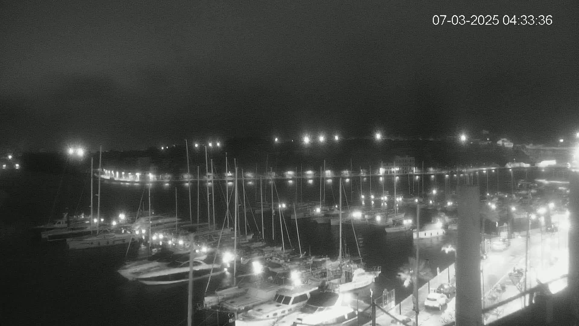 A nighttime, low-light view of a harbor filled with many boats and yachts, illuminated by lights along the docks and on the boats themselves.