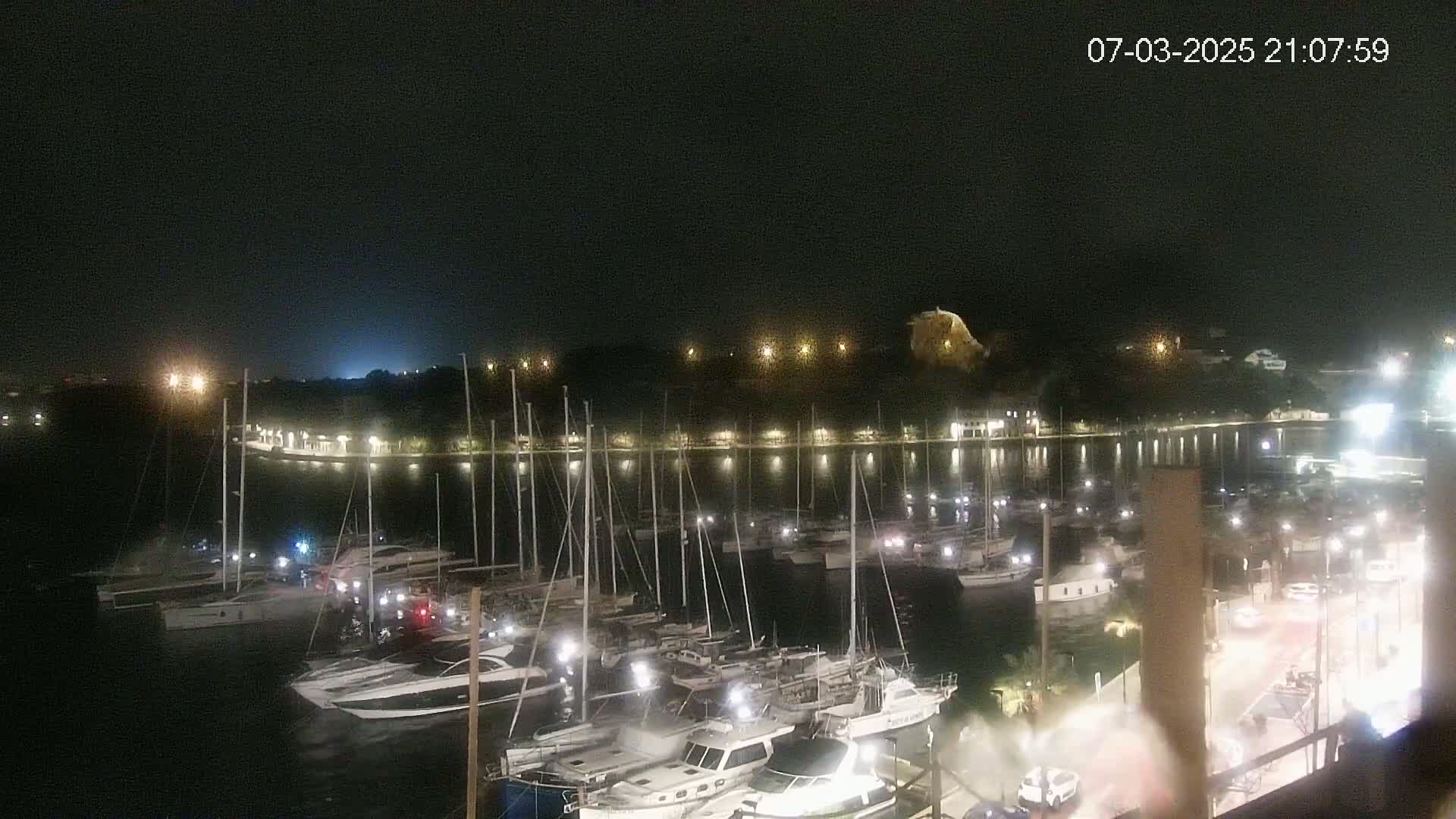 A nighttime view of a harbor filled with sailboats and motorboats, illuminated by lights on land, under a dark sky.