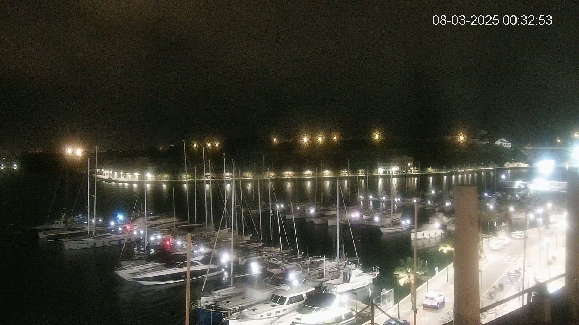A nighttime view of a marina filled with sailboats and motorboats, illuminated by lights along a waterfront promenade.