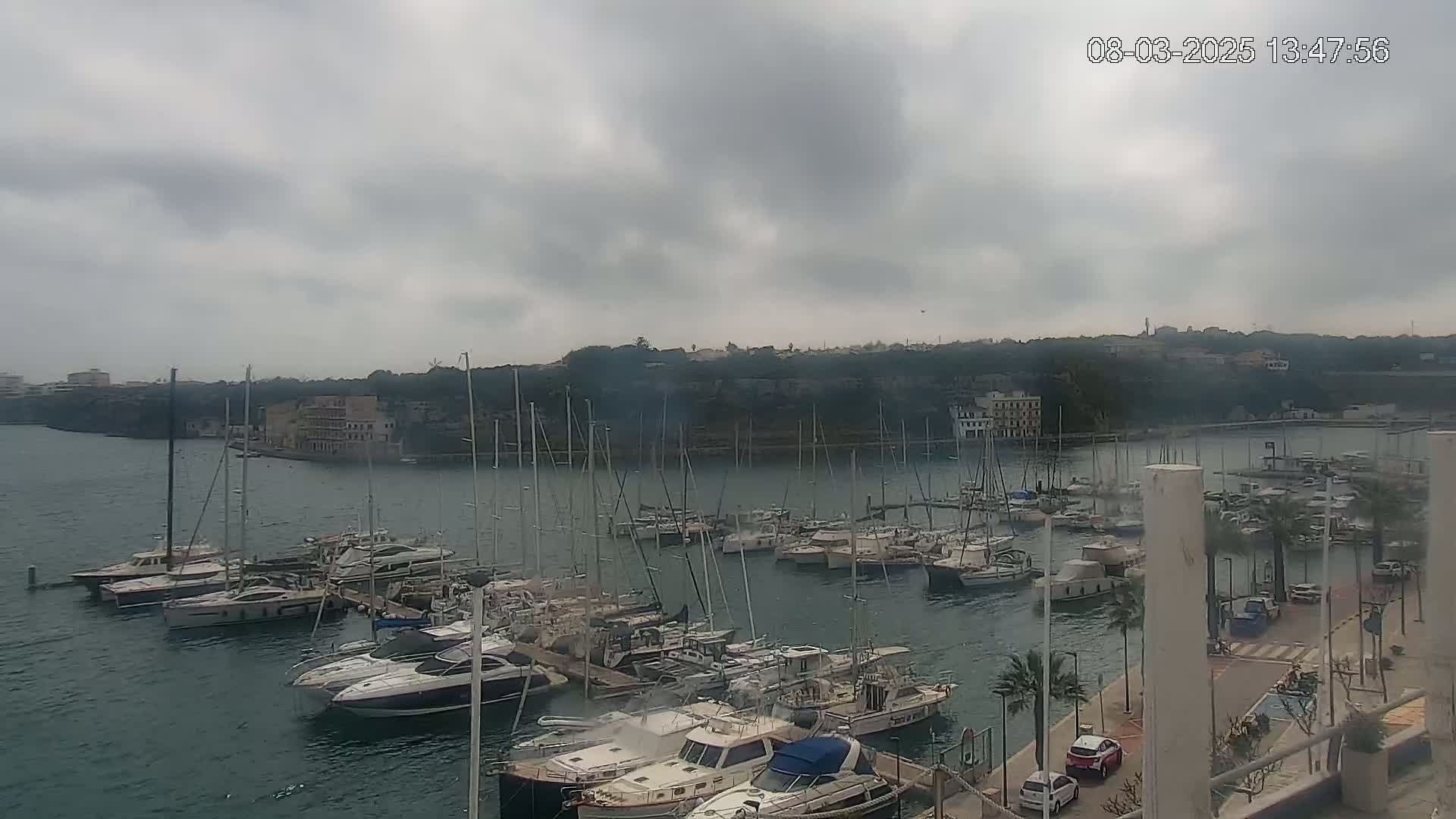 A marina filled with numerous sailboats and motorboats is situated along a rocky coastline under an overcast sky.