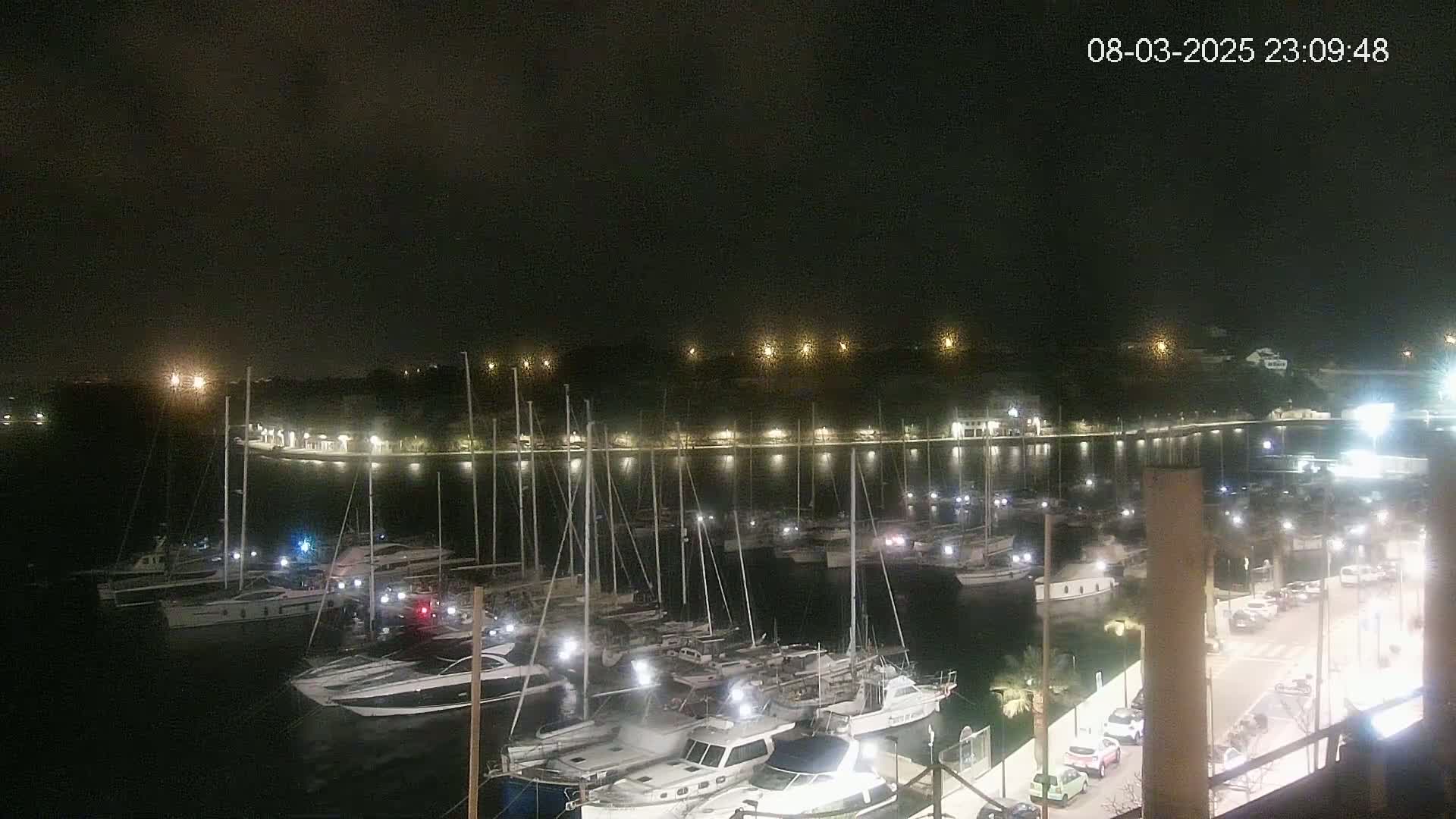 A nighttime view of a marina filled with numerous boats, illuminated by lights along a waterfront with cars parked nearby under a dark, cloudy sky.