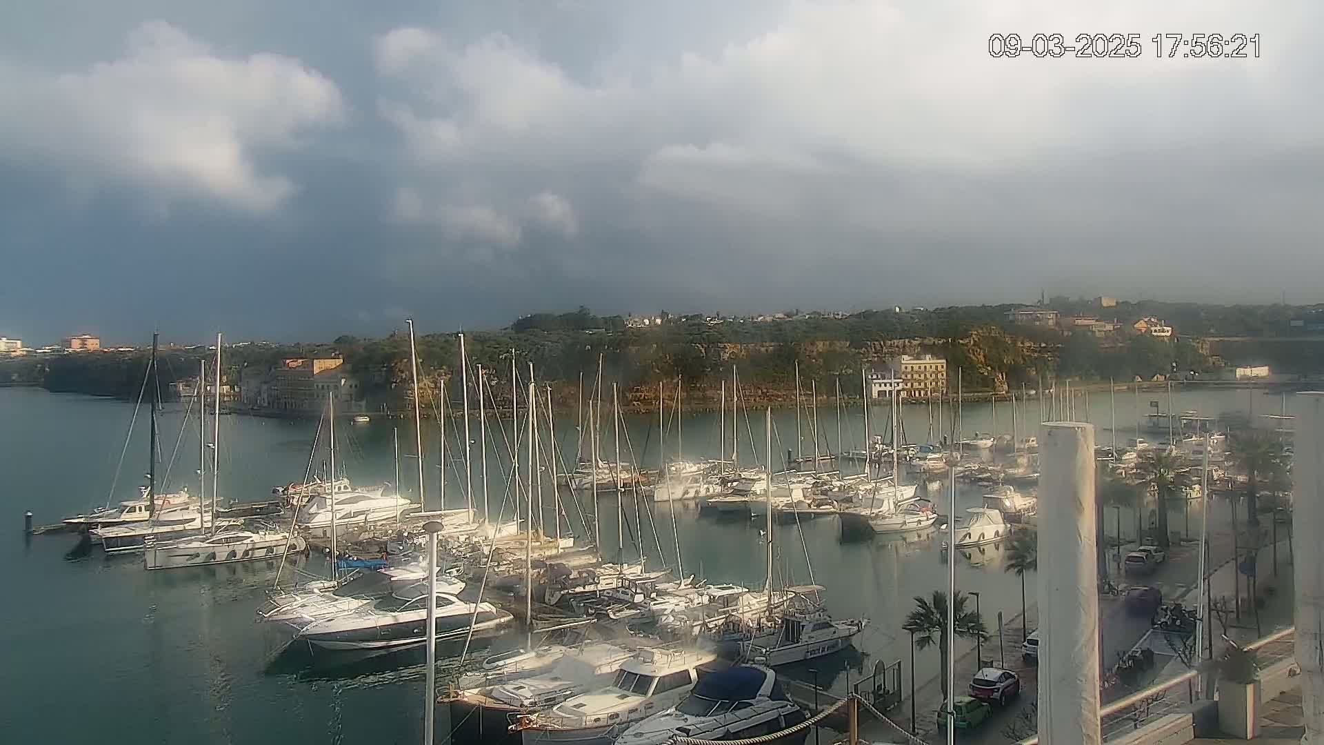 Many sailboats and motorboats are docked in a calm harbor under a partly cloudy sky.