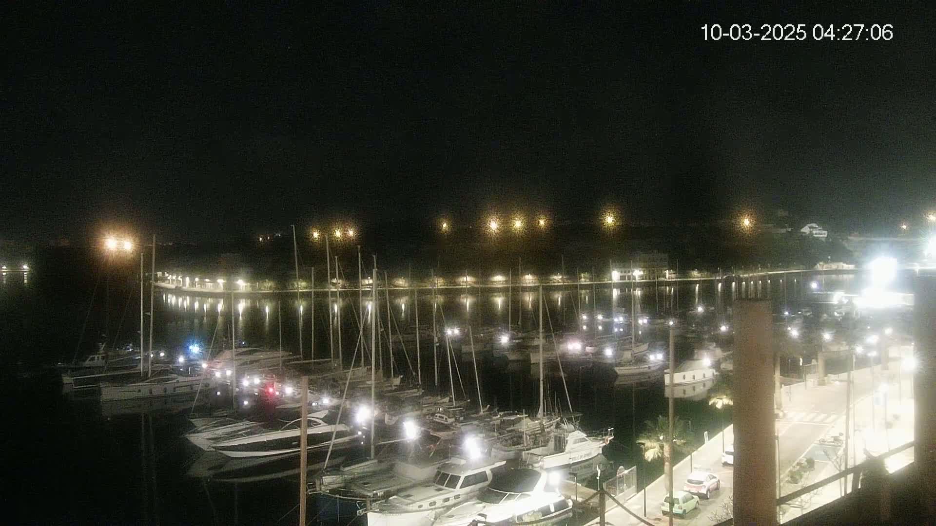 A nighttime view of a marina filled with boats, illuminated by lights along a waterfront road.