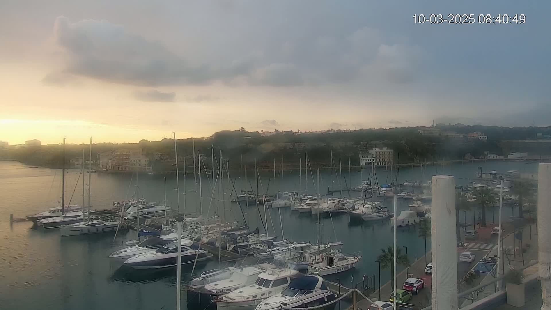 A marina filled with numerous sailboats and motorboats is seen at sunrise under a partly cloudy sky.