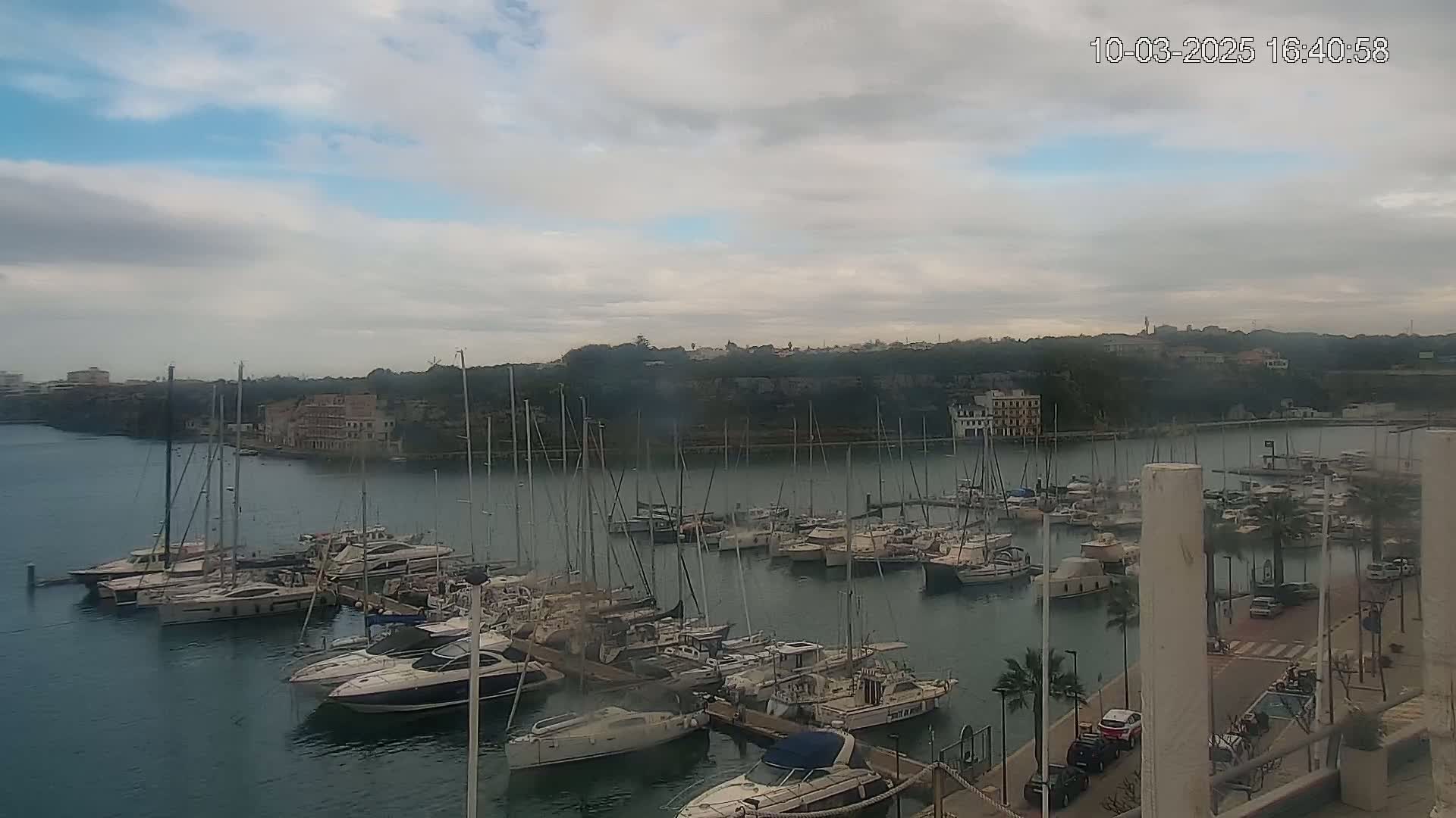 A marina filled with sailboats and motorboats is situated on calm water under an overcast sky.