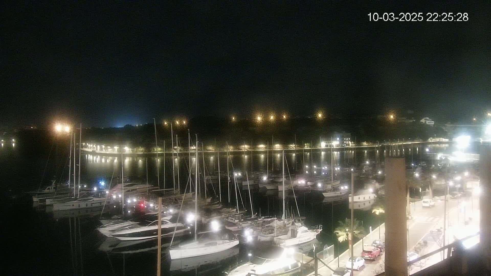 A nighttime view of a marina filled with boats, illuminated by lights along the waterfront and on the boats themselves, under a clear, dark sky.