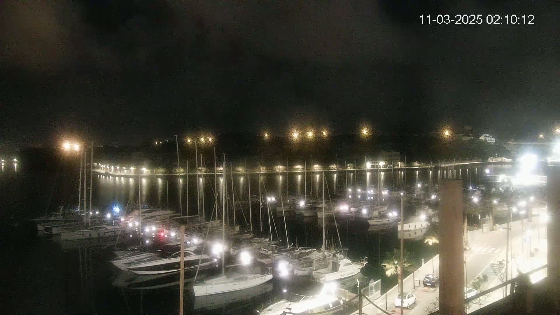 A nighttime view of a marina filled with boats, illuminated by lights along the waterfront under a dark, cloudy sky.