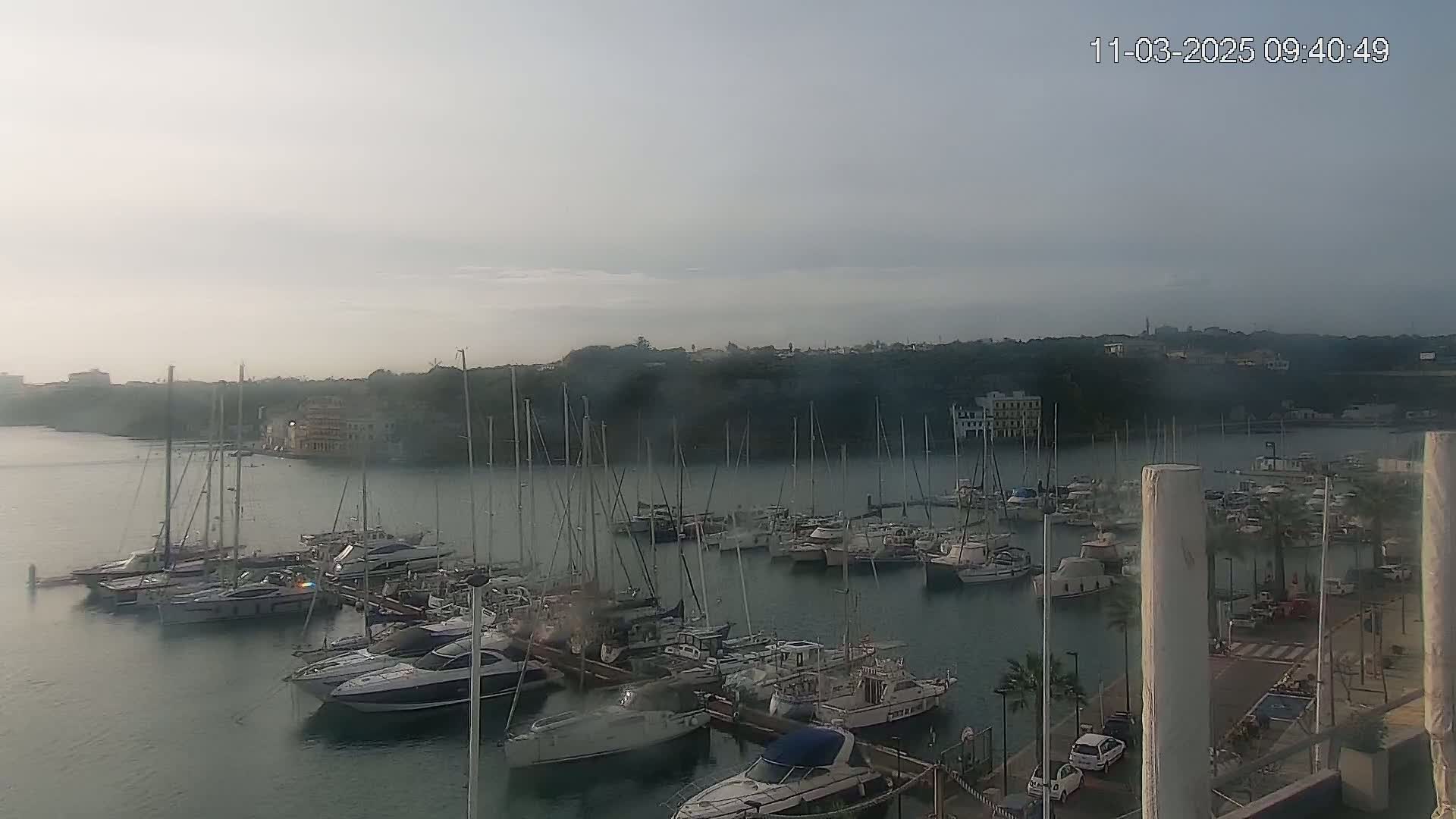 A hazy marina filled with numerous sailboats and motorboats is seen on a calm, overcast day.