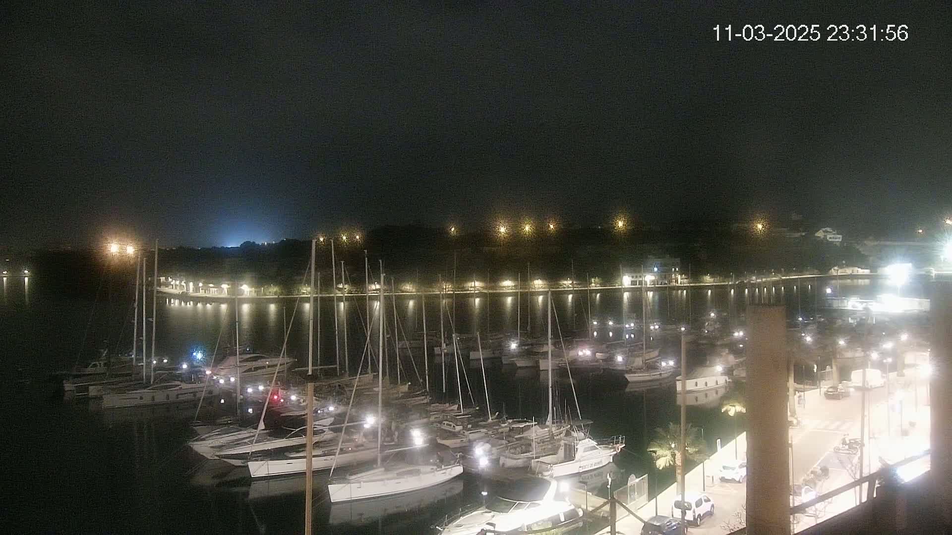 A nighttime view of a marina filled with sailboats and yachts, illuminated by lights, under a dark and clear sky.