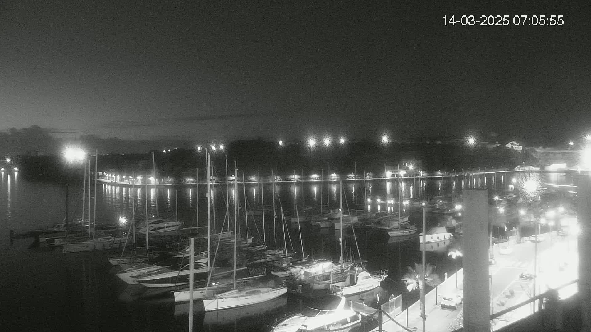 A nighttime, black-and-white view of a marina filled with sailboats and motorboats, illuminated by lights along a waterfront promenade.