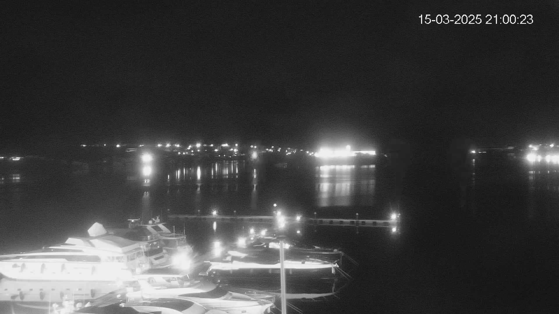 A nighttime view of a marina with several boats docked, illuminated by lights, across a calm body of water reflecting city lights in the distance.