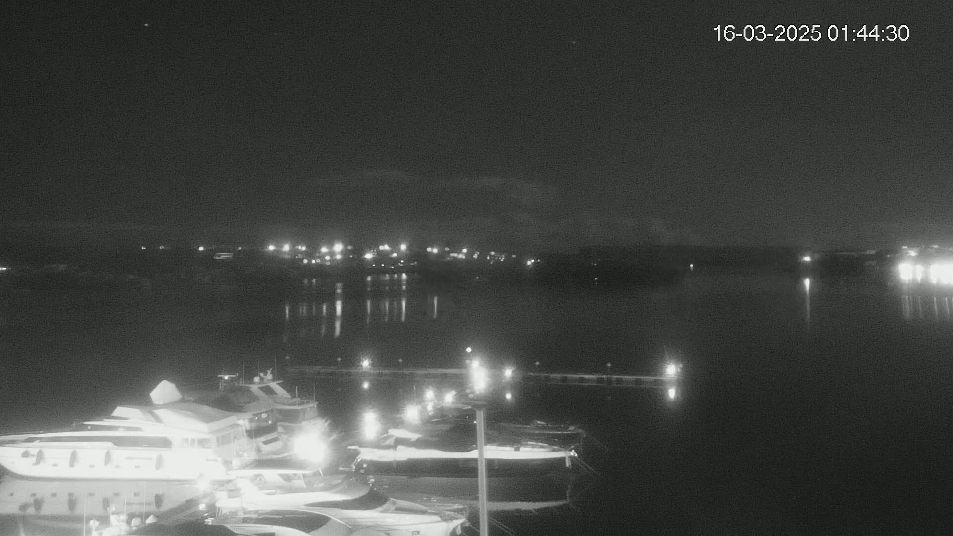 A nighttime view of a marina with several yachts docked, under a clear, dark sky, across a calm body of water from a dimly lit shoreline.