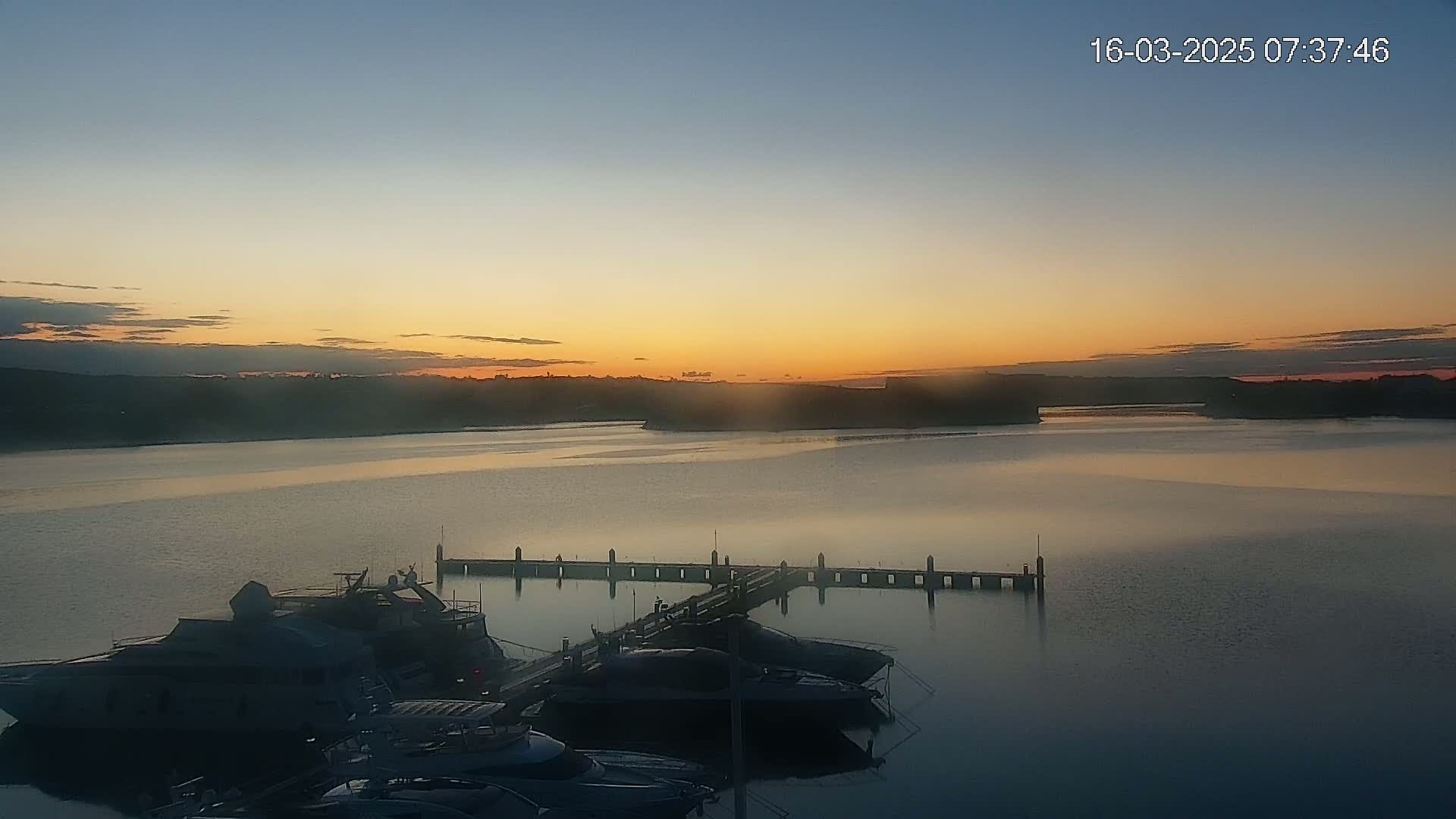 Several boats are moored at a dock during a calm sunrise over a wide body of water.