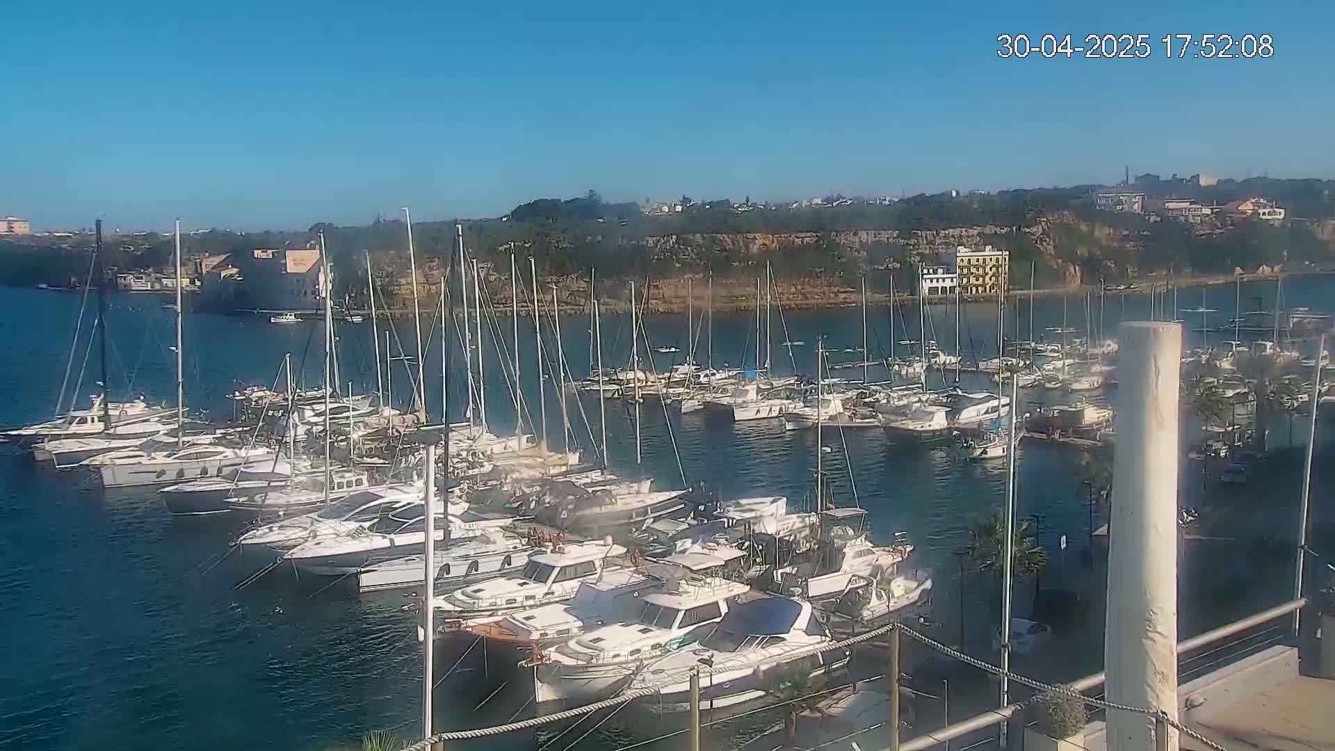 A marina filled with many sailboats and motorboats is situated along a rocky coastline under a clear blue sky.