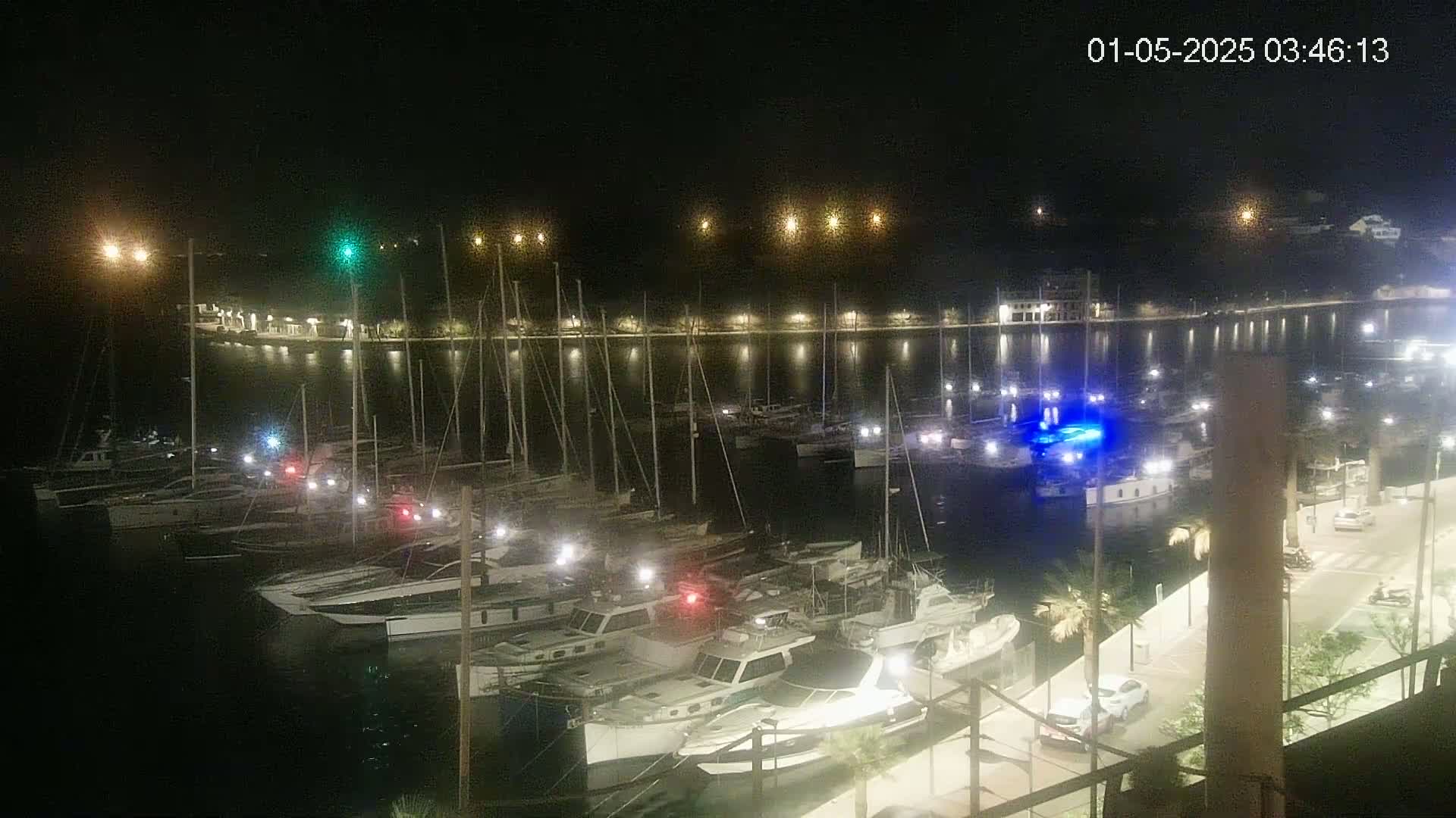 A nighttime view of a marina filled with boats, illuminated by lights along a waterfront walkway and buildings beyond.