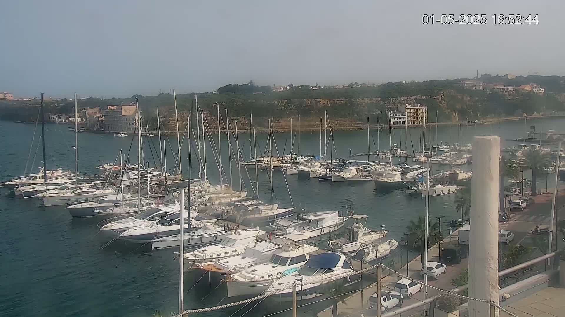 A marina filled with numerous sailboats and motorboats is situated on calm, clear water under a hazy sky.