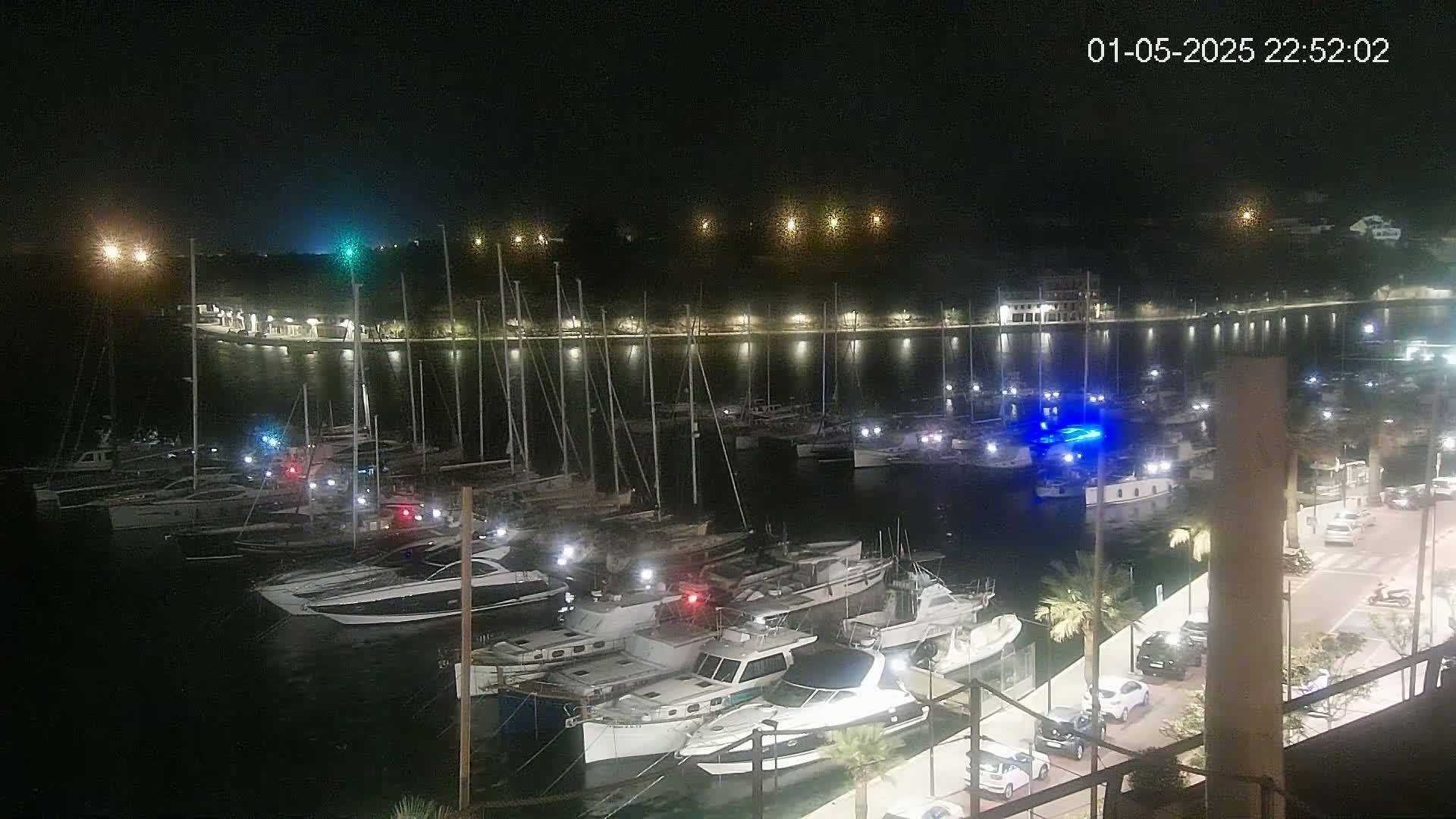 A marina at night, filled with numerous boats and yachts, is illuminated by lights along the shoreline under a clear, dark sky.