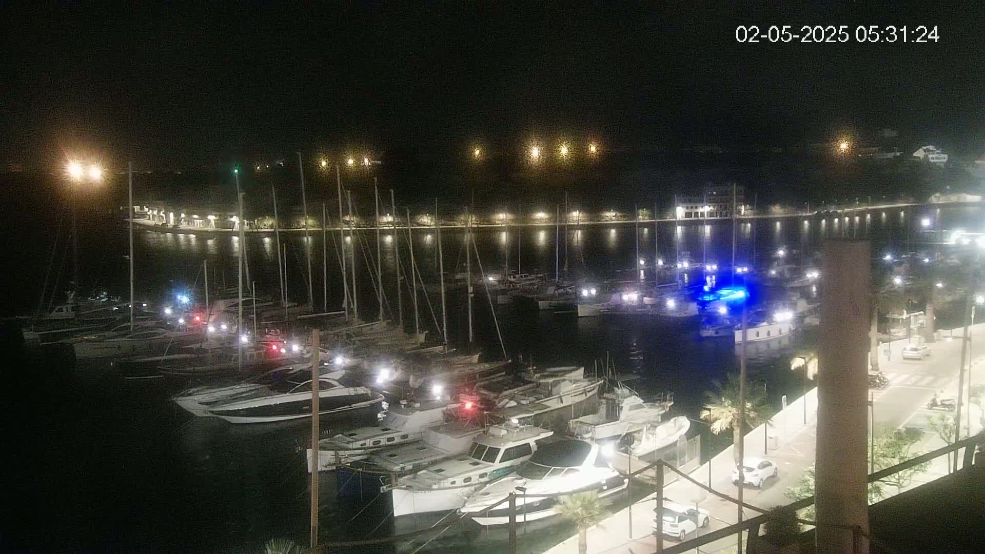Many boats are docked in a harbor at night, illuminated by lights along the shore.