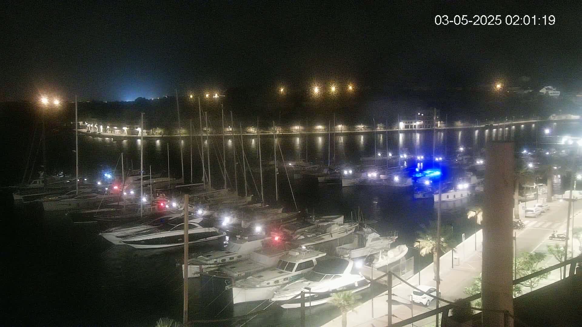 A nighttime view of a marina filled with docked boats, illuminated by lights along the waterfront and on the boats themselves.