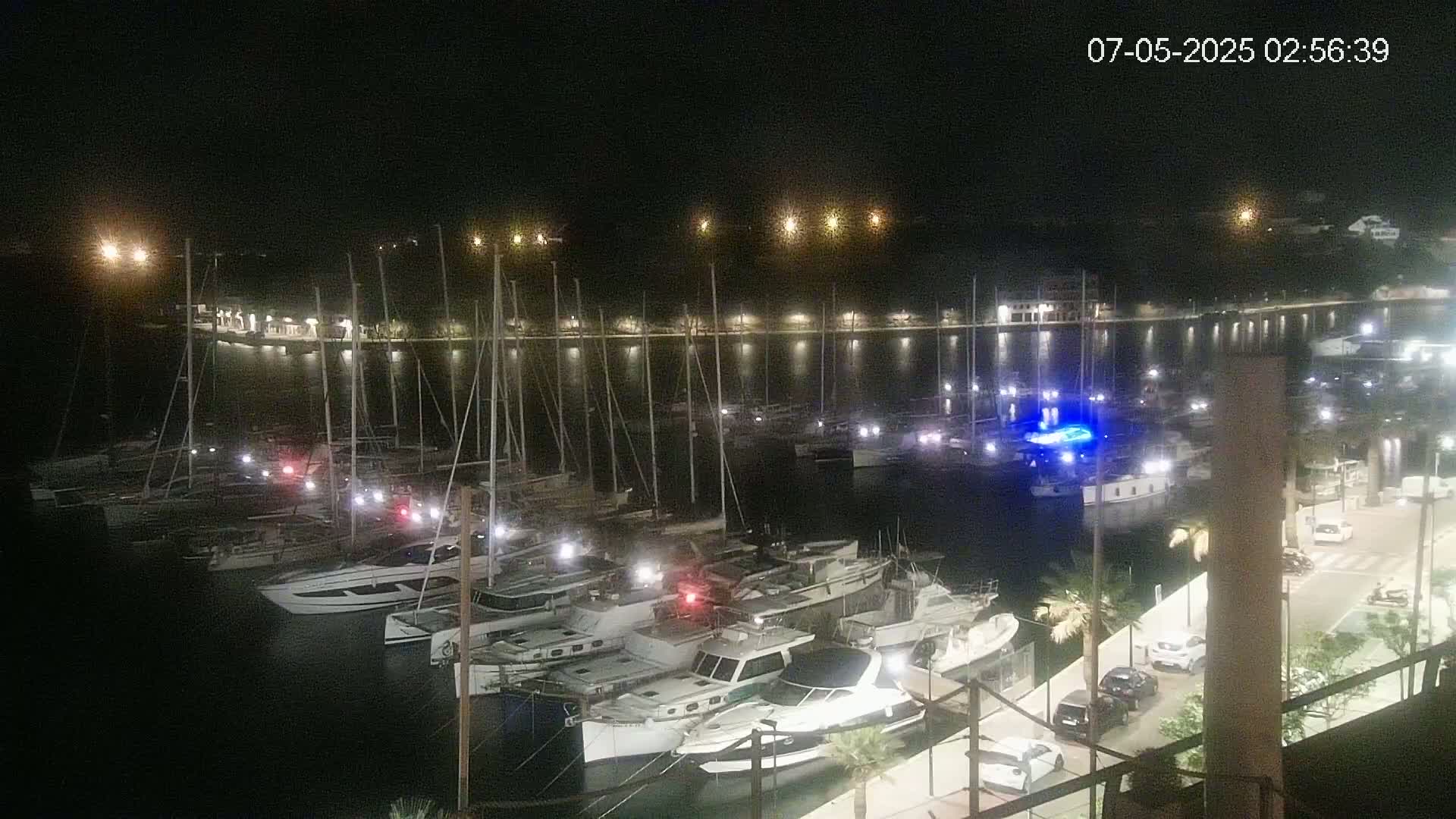 A nighttime view of a marina filled with docked sailboats and motorboats, illuminated by lights along the waterfront.