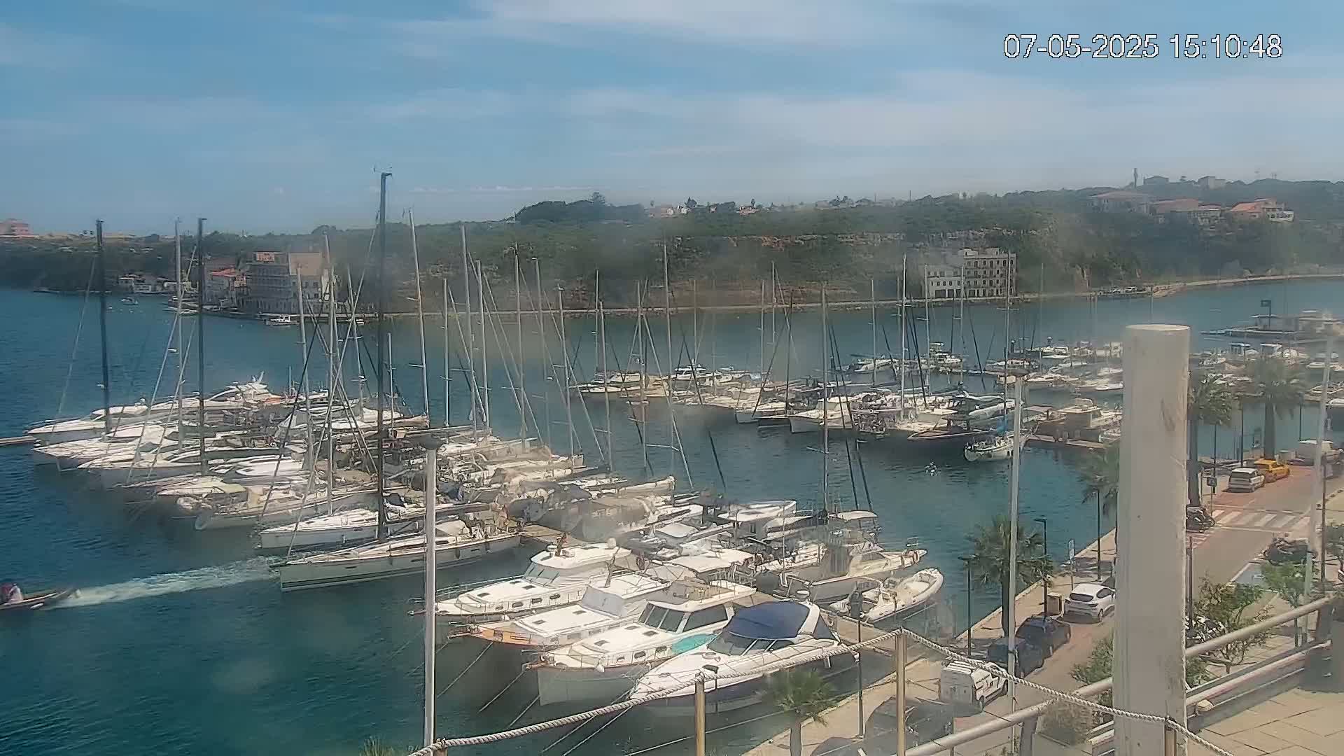 A marina filled with numerous sailboats and motorboats on a sunny day, with a small boat passing in the foreground and a shoreline with buildings and vegetation in the background.