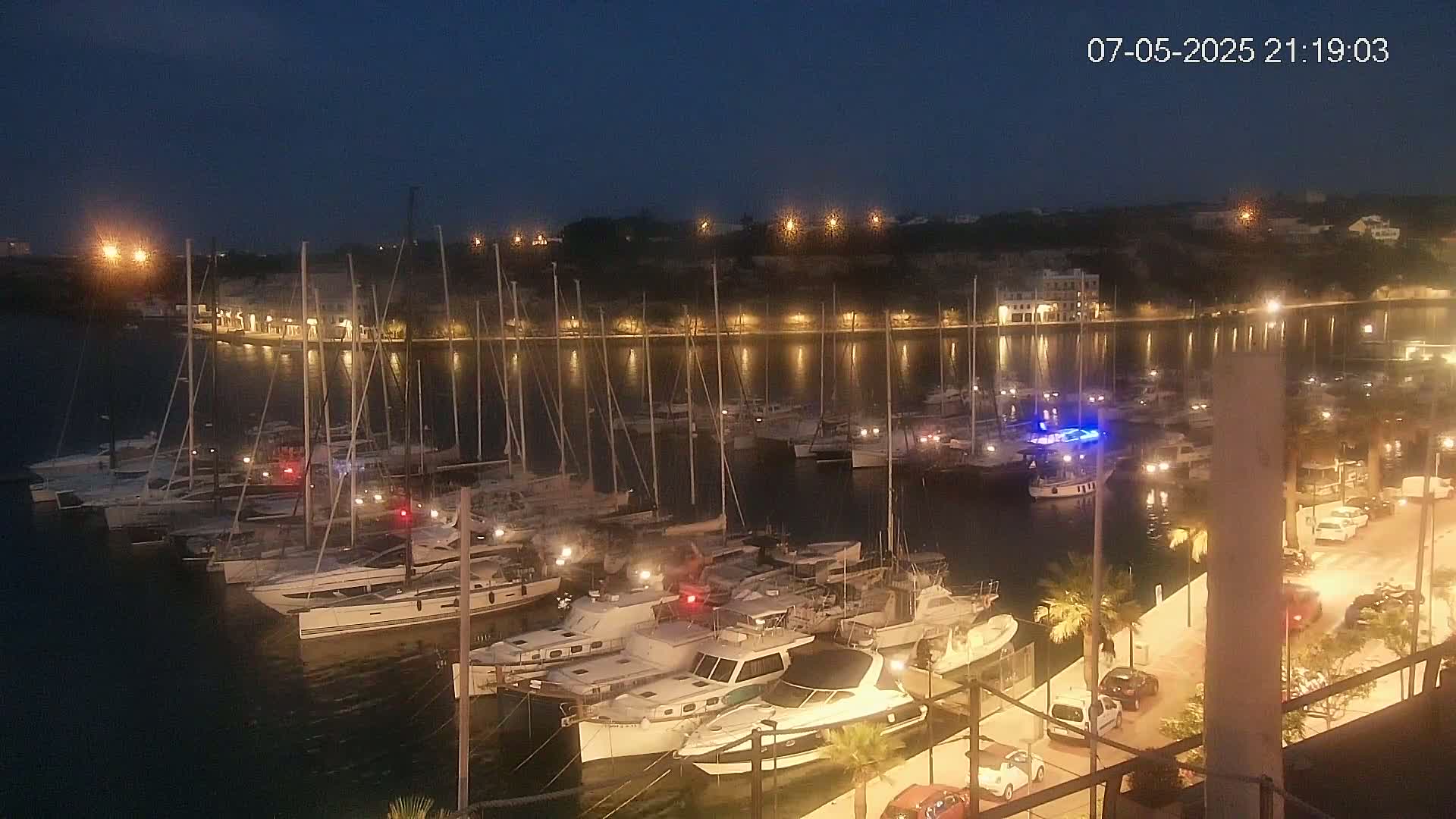 A nighttime view of a marina filled with sailboats and motorboats, illuminated by lights along a waterfront with cars parked nearby under a dark, clear sky.