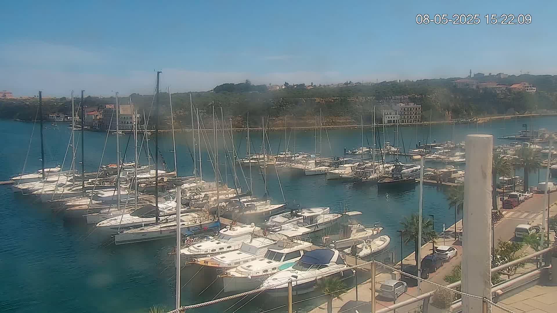 A marina filled with numerous sailboats and motorboats is seen on a sunny day, with a waterfront road and buildings visible in the background.