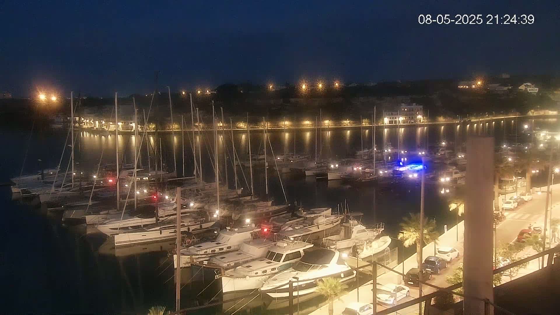A nighttime view of a marina filled with numerous boats and yachts, illuminated by lights along a waterfront promenade with parked cars.