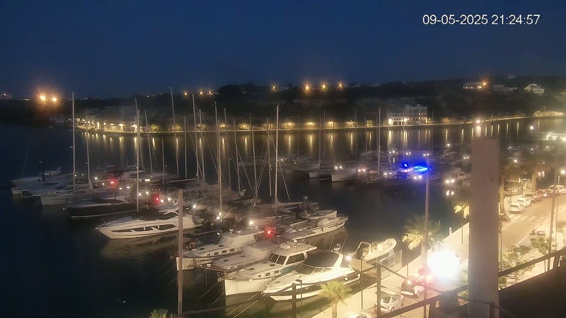 Many sailboats and motorboats are docked at a marina at night, under a dark, clear sky, with lights illuminating the waterfront.