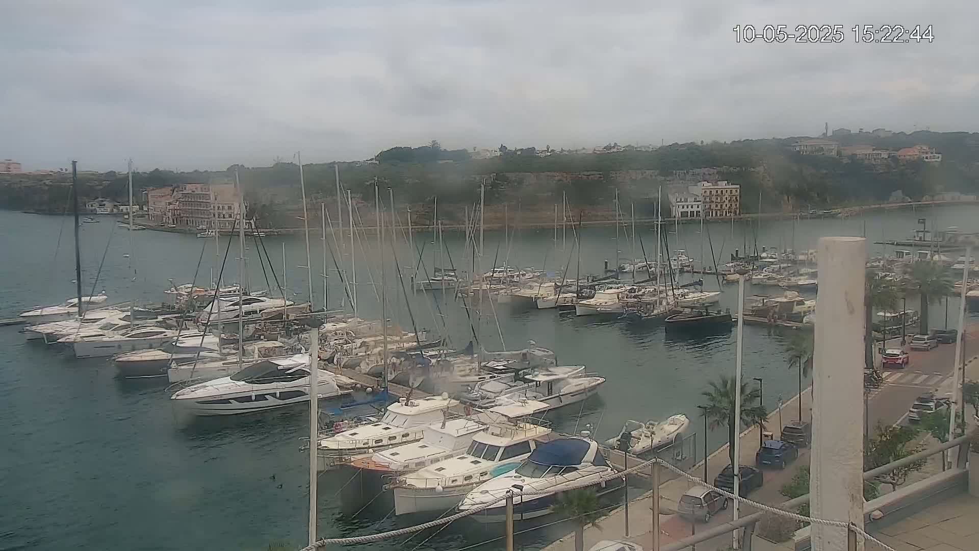 A marina filled with numerous boats and yachts is situated near a waterfront with buildings on a cloudy day.