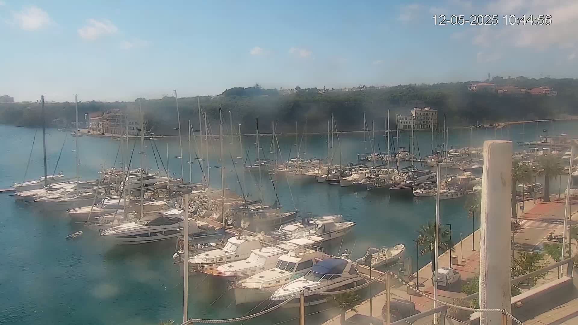 A marina filled with numerous yachts and sailboats is situated on calm, clear water under a mostly sunny sky.