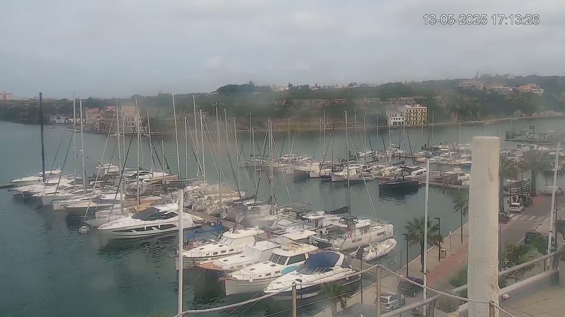 A marina filled with numerous sailboats and motorboats is situated on calm, overcast water, backed by a hilly landscape with buildings.