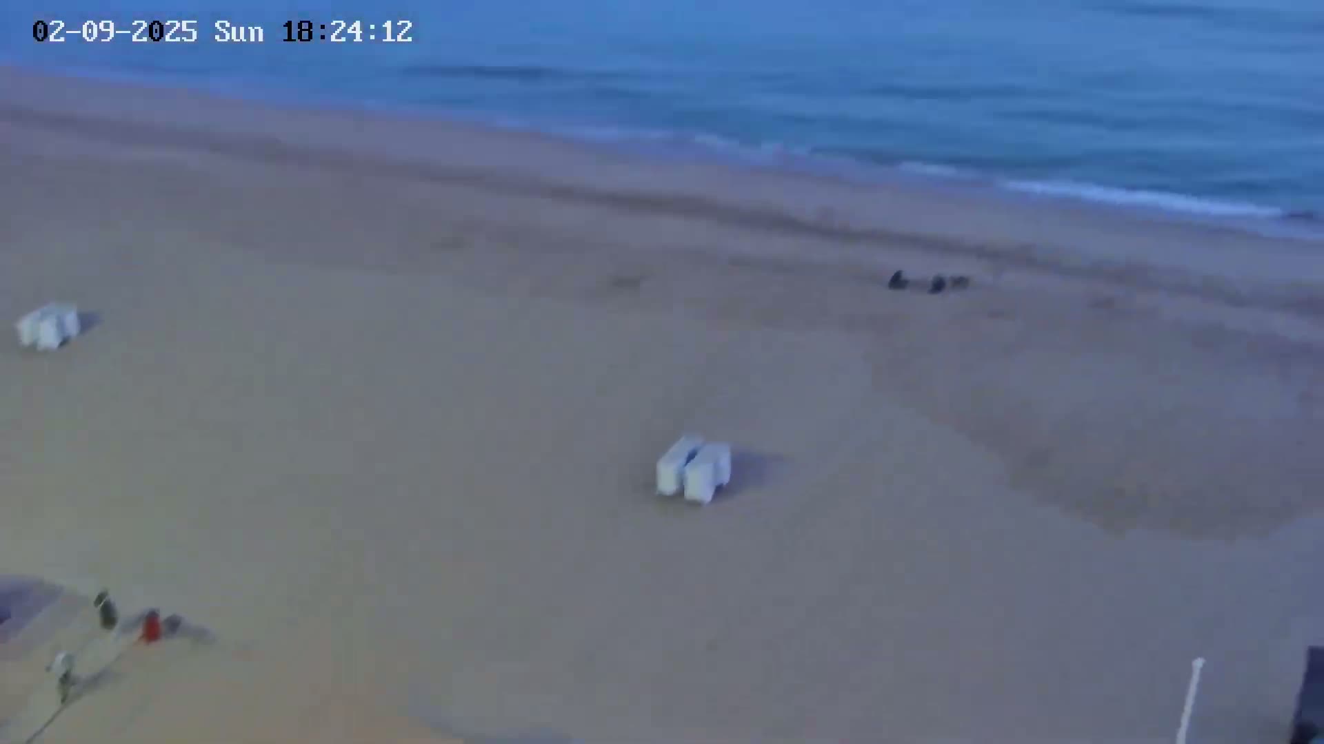 A mostly empty sandy beach is seen next to a calm, dark-blue ocean under what appears to be an overcast sky.