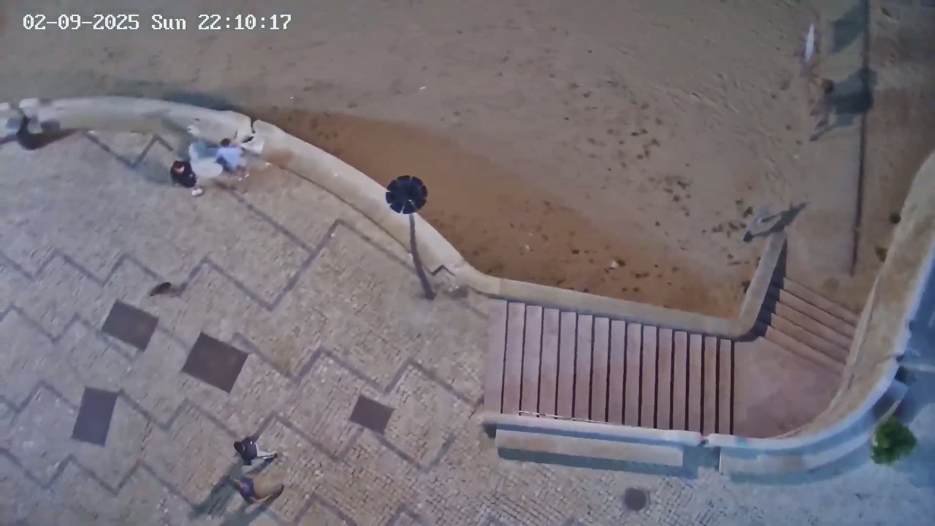 An overhead nighttime view shows several people near a sandy beach and stone steps leading up to a patterned stone walkway.