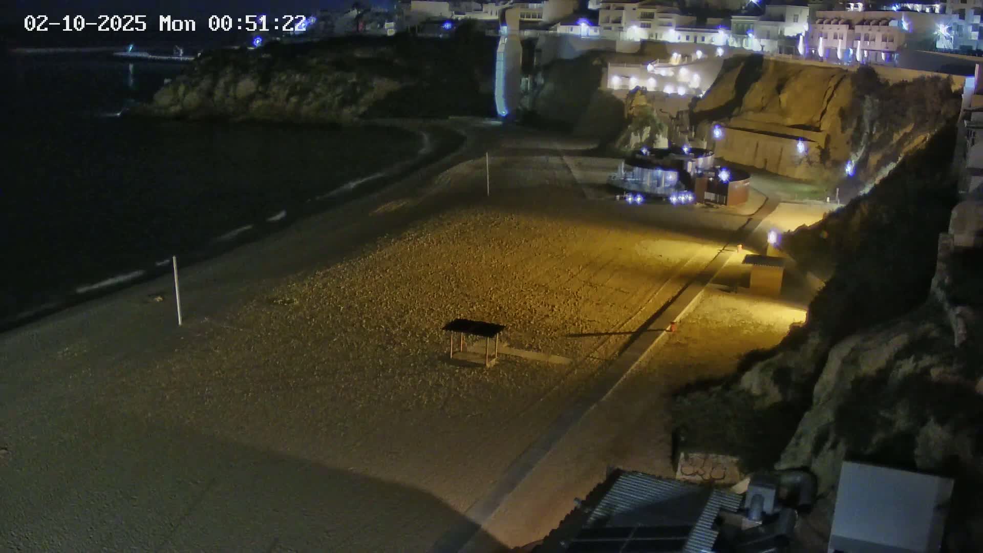 A mostly empty, pebble beach at night is illuminated by artificial lights, next to a cliffside with buildings on top.