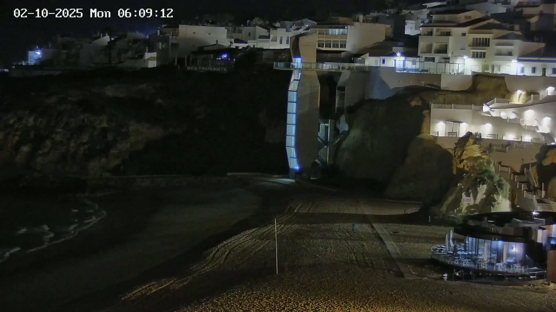 A nighttime view of a pebble beach below illuminated buildings on a cliffside, with gentle waves lapping the shore.
