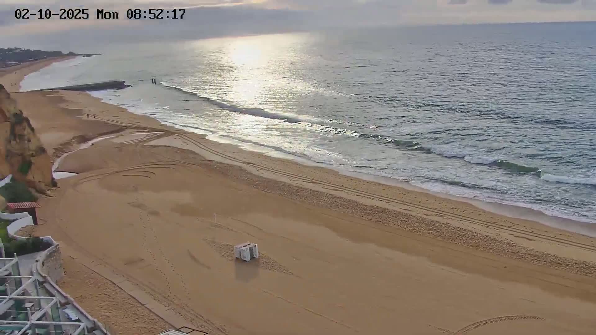 An expansive sandy beach with tire tracks stretches along a calm ocean under a partly cloudy sky.