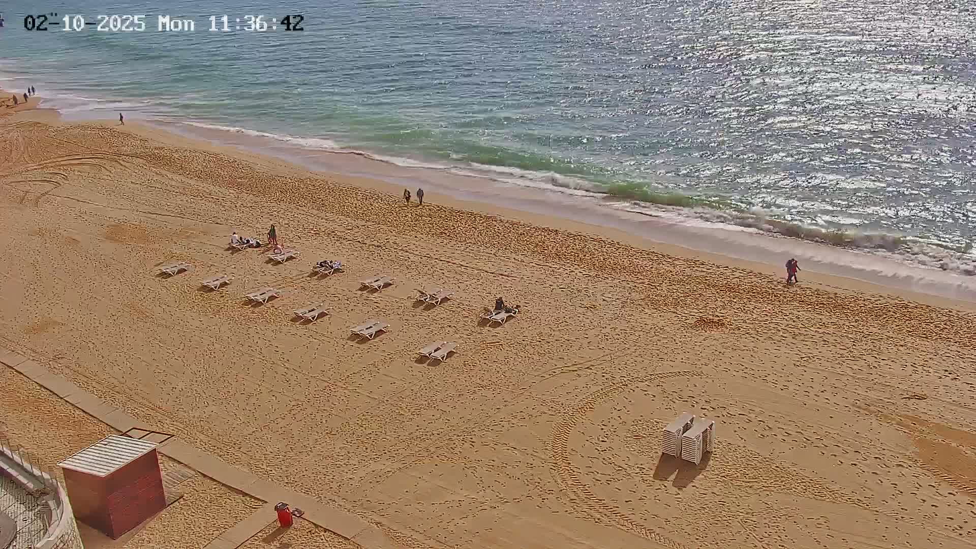 A sandy beach with a few people and lounge chairs, next to a calm, sunny ocean.