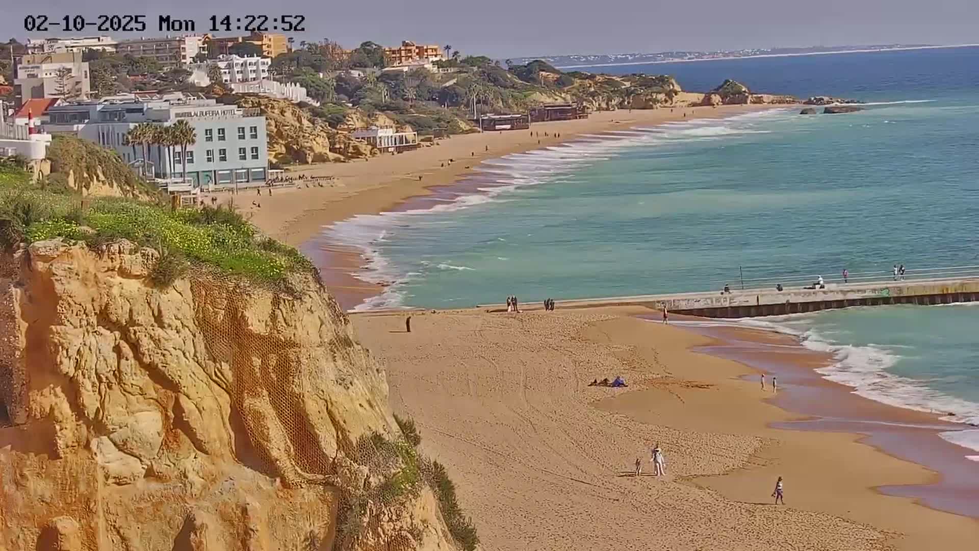 A sandy beach with turquoise water and small waves stretches beneath a cliff on a sunny day, with a few people visible on the beach and a pier extending into the ocean.