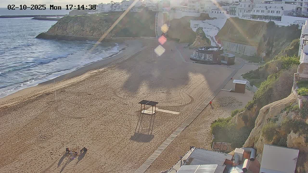 A sandy beach with a lifeguard stand and a boardwalk next to a rocky cliff overlooking the ocean on a sunny day with some small waves.